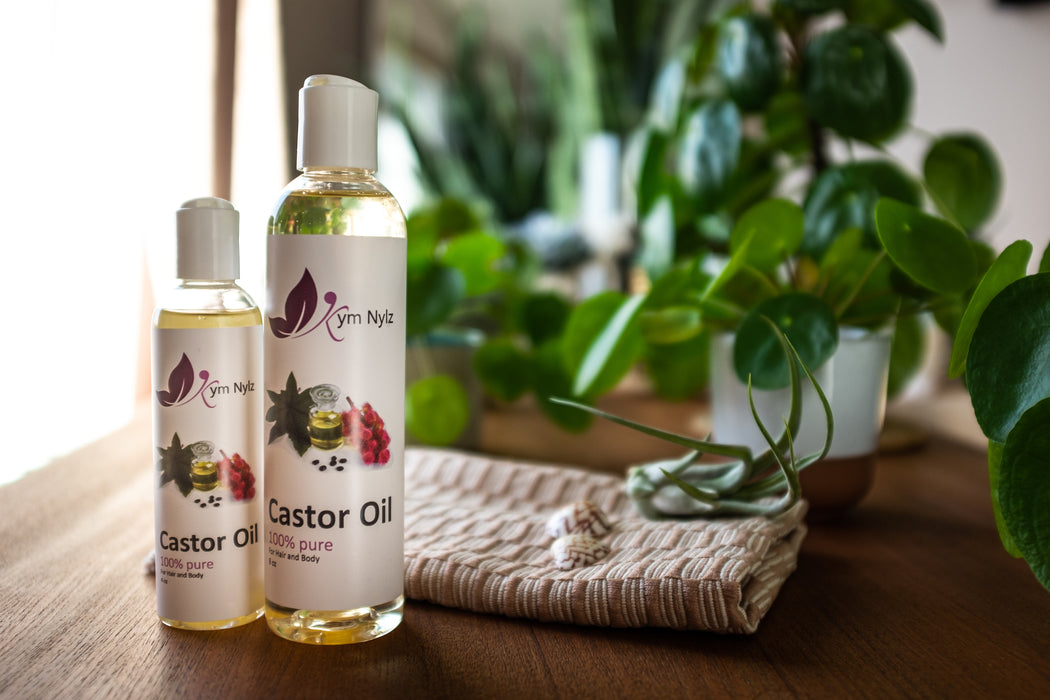 Two bottles of Kym Nylz Castor Oil - four ounce and eight ounce sizes. Sitting on a wooden desk with plants in the background.