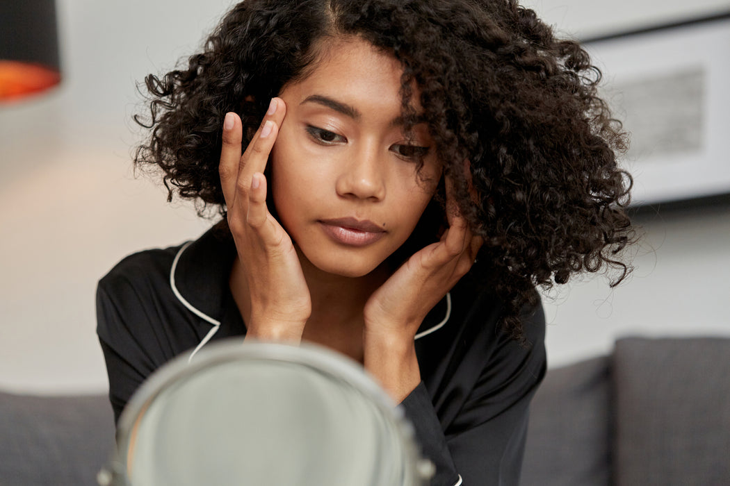 Young woman applying Kym Nylz Castor Oil to her temples while looking in the mirror. Her natural, curly, chin length hair is down, and she's wearing black pajamas.
